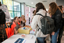 Several people are standing by a table in a bright hallway. On the table is a yellow box with materials and a bowl with a red and white pattern. Behind the table, two people in orange jackets are sitting and talking to visitors. Information posters can be seen in the background.