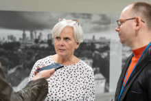 A woman with short white hair and glasses on her head, wearing a white shirt with black and red dots, is standing next to a man with glasses, dressed in an orange T-shirt and a dark blazer. They are speaking into a voice recorder.
