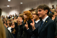 Grupa młodych osób, studentów pierwszego roku, stoi i wygłasza przysięgę studencką. Mają podniesione prawe dłonie i wyciągnięte z niej ku górze wa palce.