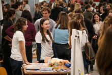 Several people are standing at a table in a crowded hallway. On the table lies a light-colored infant phantom, several small containers, and leaflets. Two people in white shirts are talking to visitors. In the background, groups of people can be seen at other stands.