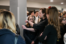 Several people are standing in the hallway. One person in a black shirt is pointing at something with their hand, and another in a dark jacket is looking at the gesture. Next to them are people in light and dark clothing, some with backpacks.