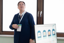 Osoba w granatowym stroju z identyfikatorem stoi przed oknem i prezentuje treści widoczne na ekranie.