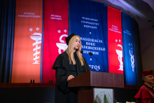 A young woman, president of the Student Council, wearing a black gown and cap, stands at the podium and gives a speech. In the background, colorful banners hang with the names of the departments and the inscription “Inauguration of the Academic Year 2025/2026.”