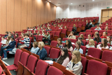 Aula wykładowa z czerwonymi fotelami, w których siedzi liczna grupa młodych osób.