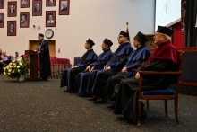 Five people in gowns and black caps are sitting on the stage. They are the associate deans and a guest lecturer. The dean of the faculty is standing at the lectern and giving a speech. Portraits hang in the background. A bouquet of white flowers lies on the floor next to the lectern.