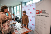 Several people are standing at a table in a bright room. There are leaflets and posters with medical graphics on the table. One person in a brown sweater is talking to a visitor in a light-colored sweatshirt. In the background, there is a white banner with a red “SOF” logo and the words “Student Onco-Forum.”