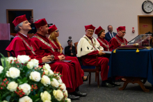 Six people in red gowns and birettas—vice-rectors, seated in the center of the auditorium during the ceremony. In the foreground on the left is a bouquet of white and red flowers. The scene depicts a moment during an academic ceremony.