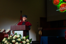A woman in a black gown and biretta stands at a lectern decorated with the university's coat of arms and a bouquet of white roses. She wears glasses. In the background, there is a screen with the inscription “Prof. Magdalena Kucia, PhD” and an image of cells under a microscope.