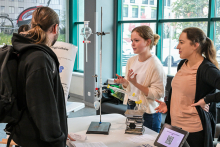 Three people are standing at a table in a bright room with large windows. On the table is a chemistry experiment kit with flasks and a stand, next to it is a tablet with a QR code and information sheets. One person in a white sweatshirt is gesturing, another in a beige T-shirt is listening, and the third in a black sweatshirt with a backpack is looking at the workstation.