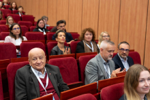 Grupa osób siedzi na czerwonych fotelach w sali konferencyjnej. Na pierwszym planie starszy mężczyzna w garniturze i czerwonym swetrze patrzy w obiektyw. Niektórzy uczestnicy klaszczą.