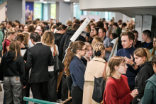 A large group of people are standing in a crowded hallway during an event. People are talking in several smaller groups, some are holding flyers. People in dark and light clothing can be seen, including suits and coats.