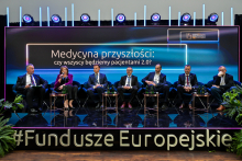Siedem osób siedzi na scenie podczas panelu dyskusyjnego. Uczestnicy noszą eleganckie ubrania i siedzą na czarnych fotelach. Przed nimi stoją stoliki z wodą mineralną. W tle świeci duży ekran z napisem „Medycyna przyszłości: czy wszyscy będziemy pacjentami 2.0?” oraz hashtagiem „#FunduszeEuropejskie”.