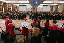 Na pierwszym planie stoi sześć osób w bordowych togach, w tym jedna w białym płaszczu z czarnymi elementami i ozdobnym łańcuchem. Przed nimi znajduje się mównica i bukiety z żółtymi kwiatami. W tle widać kilkaset osób w czarnych togach i biretach, ustawionych w rzędach, część z nich klaszcze.