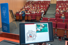 Sala wykładowa z widownią. Na ekranie komputera widoczne są ilustracje dotyczące defibrylacji. W tle osoba prowadząca prezentację.