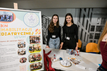 Two people are standing behind a table in a bright room. On the table there are leaflets, pens, ID badges, and a bowl with lottery tickets. Next to them there is a red plush mascot. On the left is a large poster with the words “SKN Rehabilitation Pediatrycznej WUM” (WUM Pediatric Rehabilitation Student Research Group) and group photos. In the background, there is a black-and-white graphic of a building and several orange chairs.