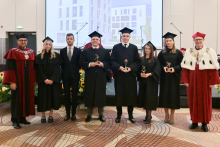 Grupa ośmiu osób. Cztery osoby w środku mają czarne togi i birety, trzymają złote statuetki na czarnych podstawach. Po bokach stoją dwie osoby w bordowych togach, jedna z nich ma biały płaszcz z czarnymi elementami i ozdobny srebrny łańcuch.