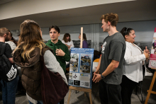 Several people are standing at a stand in a bright room. In the foreground, there is a board on an easel with a blue poster containing photos and inscriptions about a science club. One person in a green sweatshirt is talking to a person in a dark gray T-shirt, and next to them is a person in a white shirt.