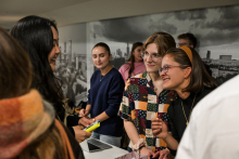 Several people are standing at a table in a bright room. In the foreground, you can see a person holding a yellow marker. There is a laptop, leaflets, and small items on the table. One person is wearing a shirt with colorful geometric patterns, and next to them is a person in a navy blue sweater. In the background, there is a black-and-white photograph of a city on the wall.