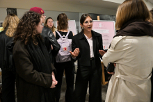 Several people are standing in a bright room at an information desk. In the foreground, there is a person in a light-colored coat holding a phone, opposite whom stands a person in a black suit with their arms outstretched. Next to them are people in dark clothes, one of whom has a light-colored backpack with a red logo. In the background, there are two posters on easels with text.