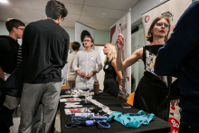 Several people are standing at a stand in a bright room. There are leaflets, a blue stethoscope, a green apron, and small medical items on the table. One person in a black dress is gesturing, and next to them are people in light and dark clothing.