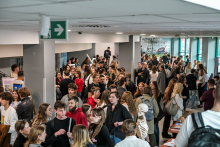 Several dozen people are standing in the hallway by the educational stands. People are talking and looking at the materials on the tables. An anatomical mannequin with a white skeleton is visible. Posters hang on the walls, and in the background there are large windows overlooking greenery.