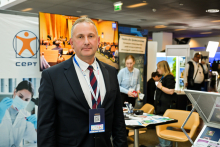 Mężczyzna, prof. Marcin Sobczak, w granatowym garniturze stoi przed stoiskiem wystawowym z logo CEPT i zdjęciem osoby w laboratorium. W tle kilka osób przegląda materiały na stołach. 