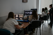  Na pierwszym planie znajduje się osoba siedząca przy biurku, patrząca na dwa monitory wyświetlające obrazy medyczne, prawdopodobnie tomografii komputerowej (CT)