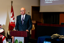 The president's advisor, dressed in a dark suit, stands behind the podium and speaks. The WUM flag can be seen in the background.