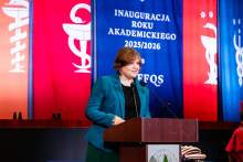 A woman, undersecretary of state at the Ministry of Science and Higher Education, wearing a green jacket, stands at the podium and speaks into the microphone. Three banners hang in the background: two red and one blue with the words “INAUGURATION OF THE 2025/2026 ACADEMIC YEAR” and the abbreviation “EFQS.” The banners feature medical symbols.