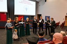 The choir during their performance. There are eight people, dressed in black suits and green and black dresses. One of the people on the left is holding a small child. Behind them is a multimedia screen, and in front of them is the audience.