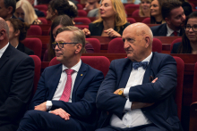 Dwie osoby siedzą w czerwonych fotelach audytorium, ubrane w eleganckie garnitury. Jedna osoba ma granatowy garnitur z białą koszulą i muszką, druga nosi ciemny garnitur z białą koszulą i różowym krawatem w drobne wzory.