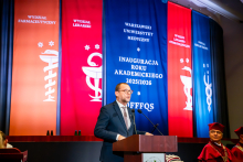 A man in a dark suit, the deputy governor of Mazovia, is speaking in the middle of the auditorium. In the background, there are colorful banners with the names of the faculties: Pharmacy, Medicine, Dentistry, and Health Sciences. On the right, there are people sitting in academic gowns.