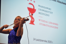 Osoba ubrana w błyszczącą, granatową suknię gra na skrzypcach, trzymając instrument pod brodą i smyczek w dłoni.