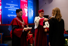Student matriculation. Two men in red gowns and birettas stand on stage. One holds a scepter, which he touches to the shoulder of the student standing in front of him.