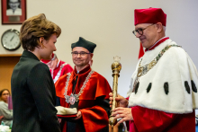 A young girl receives her student cap from the rector. Standing next to the rector is the dean of the Faculty of Pharmacy, wearing a black and orange gown.