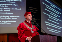 A man in a red gown and biretta, the vice-rector for science and technology transfer, stands behind the lectern and speaks. In the background, a multimedia screen displays the words of the doctoral oath.