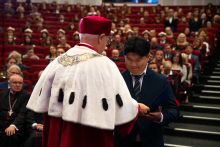 The rector presents a diploma to a man in a black suit. In the background, the audience sits in academic gowns and caps. The hall is full.