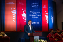 A young man, the president of the Doctoral Student Council, dressed formally, stands behind the podium and gives a speech. In the background, colorful banners hang with the names of the departments and the inscription “Inauguration of the Academic Year 2025/2026.” 
