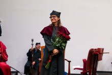 A woman in a black academic gown stands holding a bouquet of red roses. She smiles and looks to the right. In the background, a person in a red gown sits holding a scepter. 