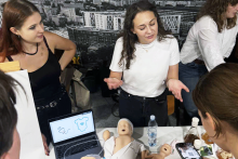 Four people are standing by a table in a bright room. On the table lies a mannequin of a baby in light-colored clothing, next to a bottle of water, leaflets, and small medical items. A laptop displays a graphic with a drawing of a hand and a T-shirt. One person in a white T-shirt is gesturing, another in a black blouse is standing next to them, and the others are leaning over the table.