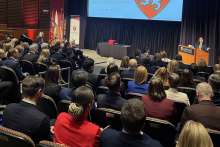 Sala audytoryjną podczas uroczystości zakończenia programu na Harvard Medical School. Na scenie stoi osoba przy mównicy z logo Harvardu i przemawia do uczestników. Uczestnicy siedzą w rzędach, a po lewej stronie stoją flagi.