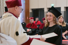 The moment of awarding the diploma. The rector, wearing a burgundy gown with a white collar, holds an open document and hands it to a young woman in a black gown and biretta, who has long, wavy hair. In the background, people in burgundy gowns are seated.