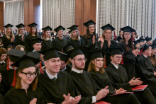 A group of people in black gowns and caps sitting in rows in a large hall with light-colored curtains and wood paneling. Several people in the middle rows are standing and applauding, and some are holding red folders on their laps.