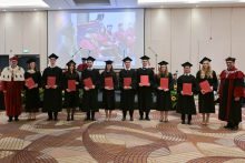 Eleven people in black gowns. Each person is holding a red folder with a diploma. On either side are two people in burgundy gowns, one of whom is wearing a white coat with black elements and a decorative chain.