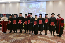Nine people in black gowns and caps hold red folders with diplomas. On either side stand two people in burgundy gowns, one of them wearing a white coat with black elements and a decorative chain.