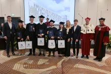 Eleven people in a large hall. Five people in black gowns and caps are holding navy blue folders. Two men are standing on either side—the rector and dean in burgundy gowns.