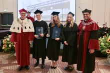 Five people standing. In the middle, three young people dressed in black gowns. Two of them are holding navy blue briefcases. On the left and right, two middle-aged men dressed in burgundy gowns. Everyone is smiling for the photo.