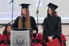 Two young women dressed in black robes stand behind a lectern. One of them is speaking. The other is holding a red briefcase. People in red robes can be seen in the background.