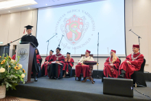 A young man in a black gown and biretta stands at a transparent lectern bearing the university's coat of arms and speaks into a microphone. In the background, six people sit in burgundy gowns, one of them wearing a white coat with black elements and a decorative silver chain.