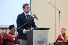 The vice president of the Regional Medical Council in Warsaw, dressed in a dark suit, stands behind the podium and speaks. Behind him, several people in red robes can be seen.